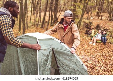 Young men pitching tent in autumn forest with blurred girls sitting on background - Shutterstock ID 619294085