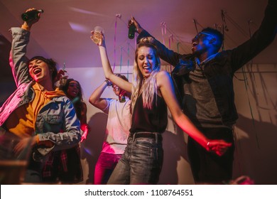 Young Men And Women Having Fun At A Colorful House Party. Friends Dancing In Joy Holding Drinks At A House Party.