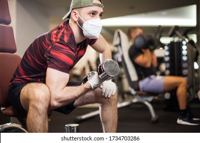 Young men working out wearing face mask latex rubber gloves,performing bicep curl with dumbbells,COVID-19 pandemic social distancing rules while working out in reopened indoor gym,prevent and protect