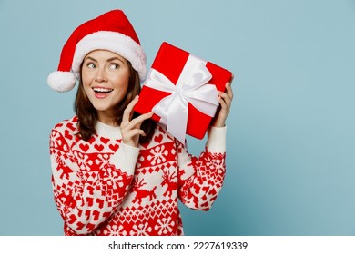 Young merry woman wear red knitted sweater Santa hat posing hold red present box with gift ribbon bow look aside isolated on plain pastel light blue cyan background Happy New Year 2023 holiday concept