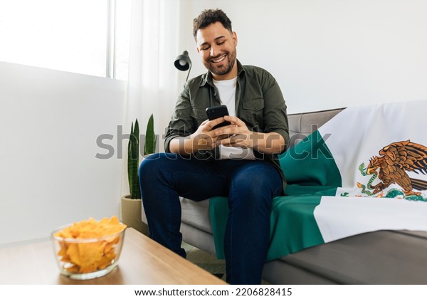 Young Mexican man using his\
cell phone to invite friends to watch a football game at his\
home.