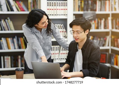 Young Mexican Professor Assisting Male Asian Student In Glasses, Helping Him With Studies In College Library