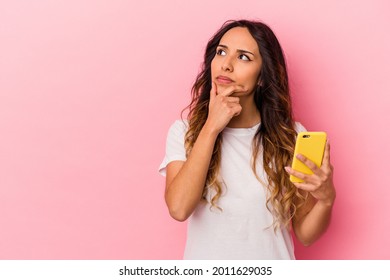 Young mexican woman holding a mobile phone isolated on pink background looking sideways with doubtful and skeptical expression.