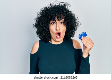 Young middle east woman playing poker holding casino chips scared and amazed with open mouth for surprise, disbelief face 
