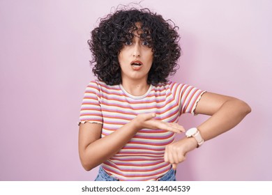 Young middle east woman standing over pink background in hurry pointing to watch time, impatience, upset and angry for deadline delay 