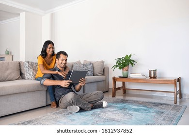 Young middle eastern couple using digital tablet while sitting on couch and floor. Happy smiling indian woman embracing from behind her boyfriend while watching video on digital tablet at home.