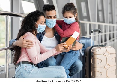 Young Middle Eastern Family Of Three Wearing Medical Masks Waiting Flight At Airport, Parents And Little Daughter Sitting With Luggage At Terminal And Checking Passports And Tickets Before Travel