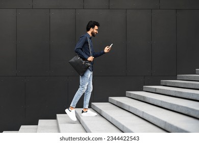 Young middle eastern guy in casual with back on his shoulder entrepreneur going up the stairs outdoor, using phone and smiling, checking email, employee scrolling on the way to office, copy space - Powered by Shutterstock