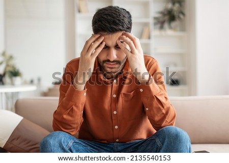 Young Middle Eastern guy suffering from headache after hard working day, sitting on couch at home, massaging head temples, having strong migraine, feeling tired, upset and stressed, copy space
