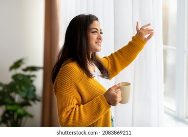 Young middle eastern lady standing by window with mug of tea, moving curtains and enjoying morning coffee in cozy living room, looking through window and smiling - Powered by Shutterstock