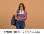 Young middle eastern lady student in a red and white plaid shirt, holding a stack of books and a backpack. She is wearing glasses and smiling at the camera. The background is a plain tan color.