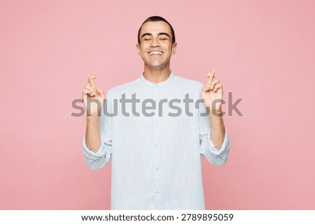 Young middle eastern man he wear casual blue cotton shirt waiting special moment, keep fingers crossed, making wish, eyes closed isolated on plain pastel light pink wall background. Lifestyle concept