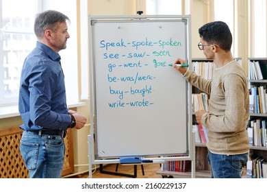 Young Middle Eastern man standing at whiteboard writing past participle forms of irregular verbs during lesson - Shutterstock ID 2160229137