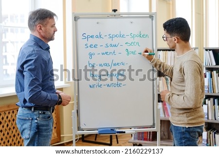 Young Middle Eastern man standing at whiteboard writing past participle forms of irregular verbs during lesson