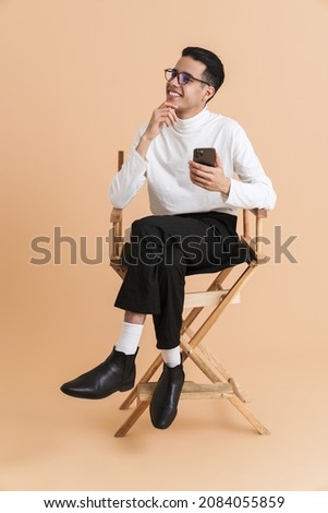 Young middle eastern man using cellphone while sitting on chair isolated over beige background