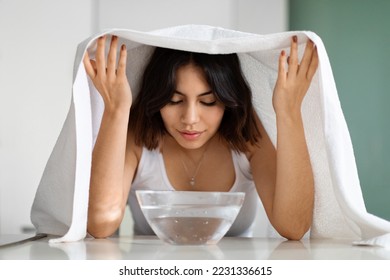 Young middle eastern short-haired woman with closed eyes sitting at table with bowl with hot water on, covered with bath towel, making facial steam beauty treatment, home interior - Shutterstock ID 2231336615