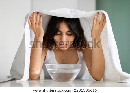 Young middle eastern short-haired woman with closed eyes sitting at table with bowl with hot water on, covered with bath towel, making facial steam beauty treatment, home interior