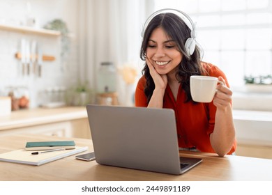 Young middle eastern woman with headphones using laptop and enjoying a cup of coffee, embodies multitasking at home - Powered by Shutterstock