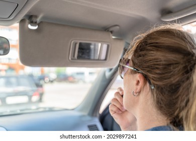 A young middle-aged woman does make-up paints her lips with lipstick in the car while looking into a small mirror in a visor with light on the go. Rear view reflected in the mirror.