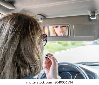 A young middle-aged woman does make-up paints her lips with lipstick in the car while looking into a small mirror in a visor with light on the go. Rear view reflected in the mirror. Selective focus.