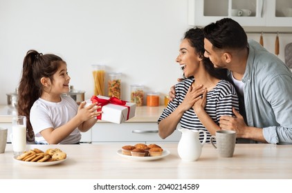 Young Middle-eastern Father And Daughter Greeting Surprised Woman With Birthday, Little Girl Giving Big Festive Gift Box While Having Breakfast Together In Kitchen. Family Birthday Celebration Concept