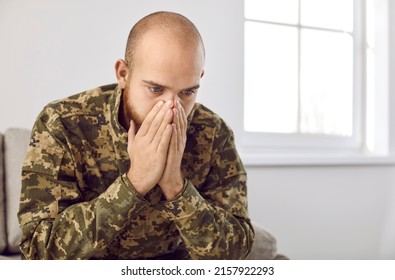 Young Military Man Suffering From PTSD. Depressed Veteran Soldier Wearing Camouflage Uniform Sitting On Couch, Holding Hands On Face And Staring Vaguely At Nothing Unable To Forget Horrors Of War