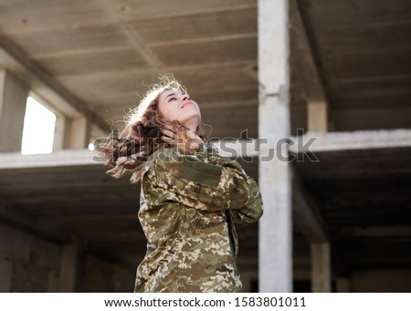 Young military woman, wearing ukrainian army military uniform and black t-shirt waving her curly blond hair in rim light. Three-quarter portrait of female soldier in front of ruined abandoned building