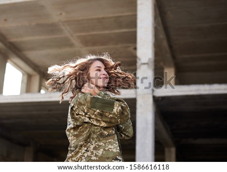 Young military woman, wearing ukrainian army military uniform and black t-shirt waving her curly blond hair in rim light. Three-quarter portrait of female soldier in front of ruined abandoned building