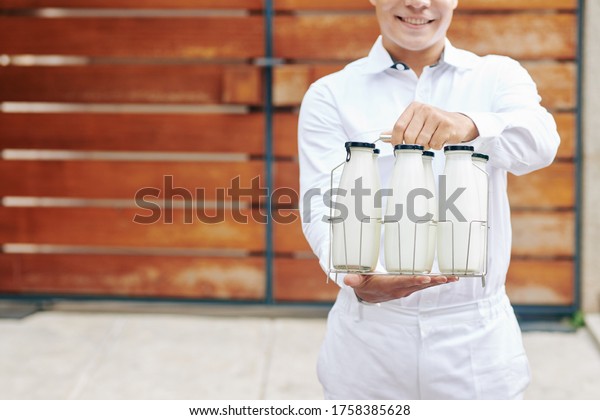 Young milkman in perfect white uniform standing\
against wooden fence holding glass bottles with milk, head cropped\
shot, copy space