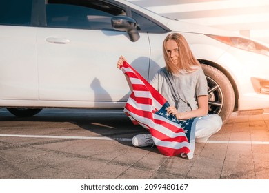 Young millennial blonde woman sitting with american flag next to car. Flag of the united states in her hands. July 4th Independence Day. USA national holiday. Freedom and memorial concept