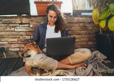 Young Millennial Hipster Girl In Casual Look Working At Sunny Home Terrace On Laptop Computer Enjoying Good Weather With Dog Lying Next To Her, People Remote Work Lifestyle