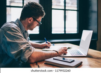 Young millennial male student searching information for homework using laptop and wifi connection indoors, man writing article in textbook for add interesting content on own website or web page