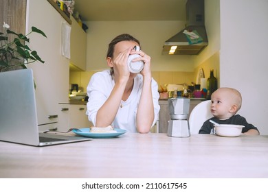 Young Millennial Mother With Coffee Fighting Tiredness While Breakfast With Baby In Kitchen. Freelancer Mom And Child After Sleepless Night. Woman Studying Or Working Online At Home On Maternity Leave