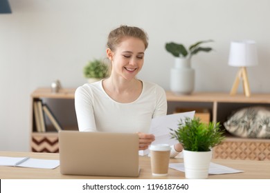 Young millennial satisfied woman sitting at the desk in office room, at home. Glad worker female holding papers report letter feels proud and happy. Smart student received good news about scholarship