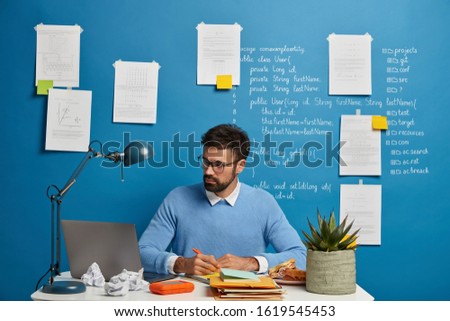 Young millennial student searches information for homework, writes down necessary content in textbook, prepare project on IT topic, sits at desktop with laptop computer, stuck papers on blue wall