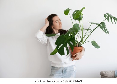 Young millennial woman holding a potted green monstera plant. Decorating apartment with plants. Home gardening hobby. - Shutterstock ID 2337061463