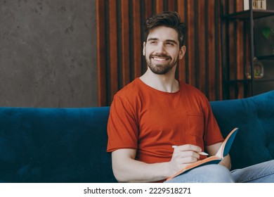 Young minded dreamful man wear red t-shirt look aside write down memories in notebook diary sit on blue sofa stay at home hotel flat rest relax spend free spare time in living room indoors grey wall - Powered by Shutterstock