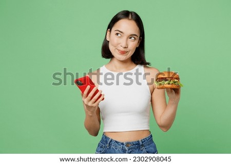 Young minded fun woman wear white clothes hold eat burger use mobile cell phone look aside isolated on plain pastel light green background. Proper nutrition healthy fast food unhealthy choice concept