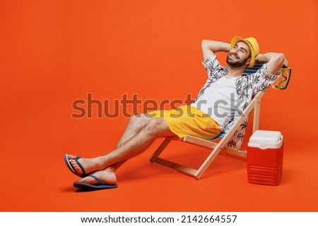 Young minded happy cool tourist man in beach shirt hat lie on deckchair near fridge hold hands behind neck isolated on plain orange background studio portrait. Summer vacation sea rest sun tan concept