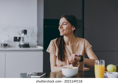 Young Minded Happy Punctual Housewife Woman 20s Wear Beige T-shirt Eat Breakfast In Morning Look Aside Use Smart Watch Check Time Cooking Food In Light Kitchen At Home Healthy Diet Lifestyle Concept