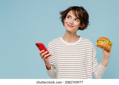 Young Minded Happy Woman In Striped Shirt Using Mobile Cell Phone Hold Burger Count Calories Browsing Internet Think Isolated On Plain Pastel Light Blue Background Studio People Lifestyle Food Concept