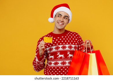 Young minded man wear red knitted christmas sweater Santa hat hold shopping paper package bag credit card look aside isolated on plain yellow background Black Friday New Year 2023 sale buy day concept