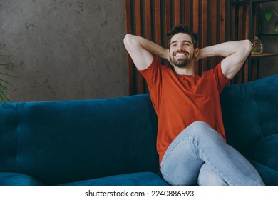 Young minded man wears red t-shirt hold hands behind neck look aside sit on blue sofa stay at home hotel flat rest relax spend free spare time in living room indoors grey wall. People lounge concept - Powered by Shutterstock