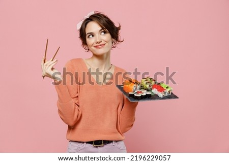 Young minded pensive thoughtful fun woman in sweater hold in hand makizushi sushi roll served on black plate traditional japanese food look aside chopsticks isolated on plain pastel pink background.