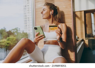 Young Minded Sportswoman Woman In White Sportswear Earphones Listen Music Use Mobile Cell Phone Credit Bank Card Shopping Online Order Delivery Book Tour Pay Gym Membership Sit On Windowsill Indoor