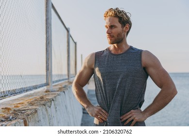 Young minded strong sporty athletic toned fit sportsman man 20s wear sports clothes warm up train stand akimbo ars on waist at sunrise sun over sea beach outdoor on pier seaside in summer day morning.