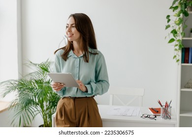 Young minded successful employee business woman 20s in blue shirt work use hold tablet pc computer look aside stand at workplace white desk at light modern office indoors. Achievement career concept