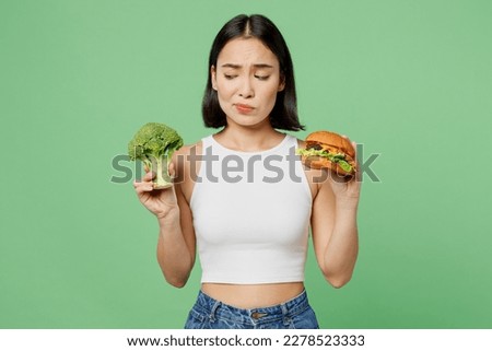 Young minded thoughtful sad woman wear white clothes hold burger broccoli count calories isolated on plain pastel light green background. Proper nutrition healthy fast food unhealthy choice concept