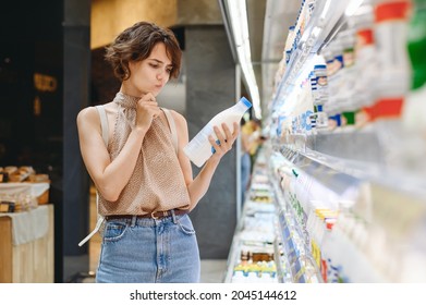 Young Minded Woman 20s In Casual Clothes Backpack Shopping At Supermaket Store Buy Choose Dairy Produce Take Milk Read Shelf Life Prop Up Chin Inside Hypermarket. Purchasing Gastronomy Food Concept.