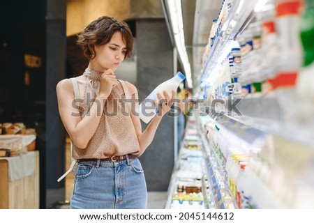 Young minded woman 20s in casual clothes backpack shopping at supermaket store buy choose dairy produce take milk read shelf life prop up chin inside hypermarket. Purchasing gastronomy food concept.